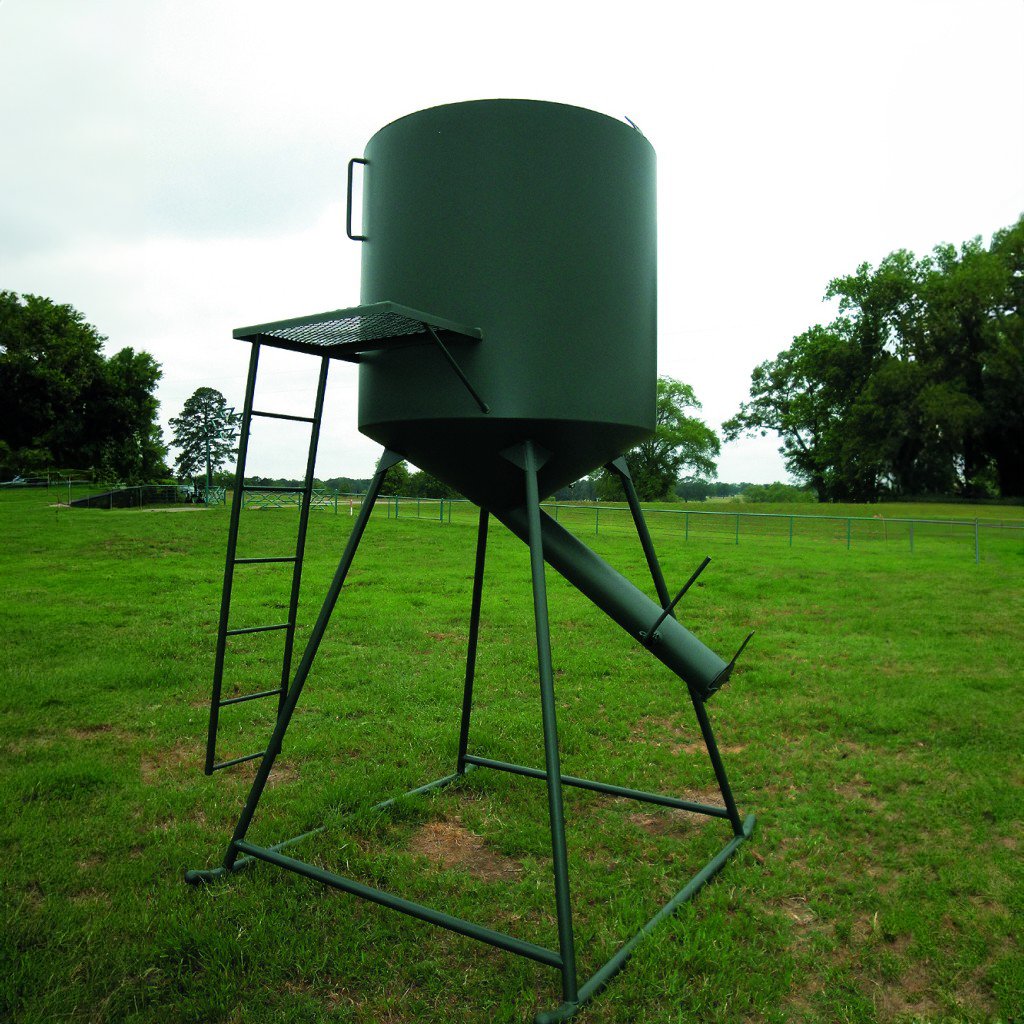 A green bulk bin feeder on a metal stand, placed on a grassy field.