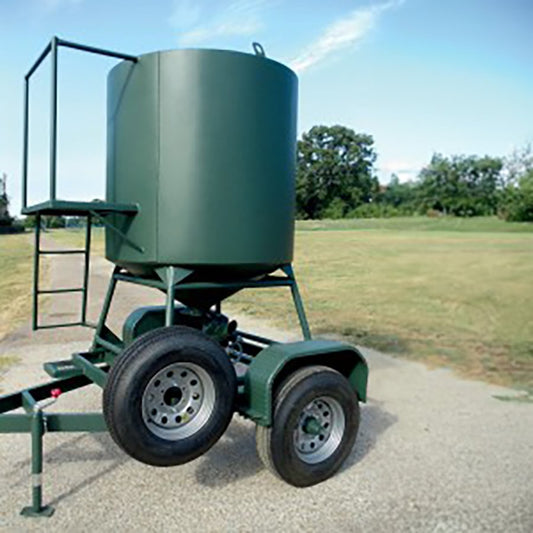A green metal quail tender trailer with a cylindrical feeder on top and wheels attached to a hitch, set against an outdoor backdrop.