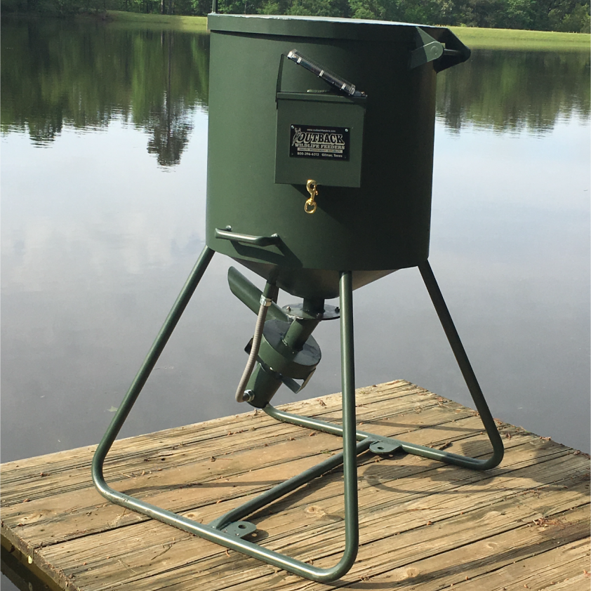 A green cylindrical fish feeder placed on a wooden dock with a body of water and trees in the background.
