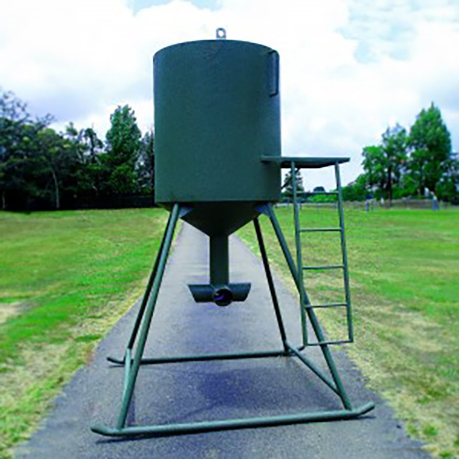 A green protein feeder mounted on a stand with a ladder, situated on a road with trees in the background.