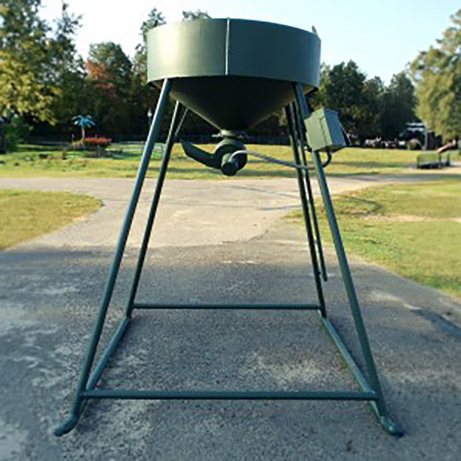 A green metal wildlife feeder with a cylindrical hopper and a rotating door attached to a stand, set on a concrete surface with trees and grass in the background.