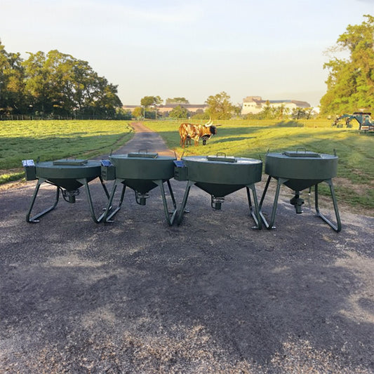 A series of four metal wildlife feeders arranged in a line on a road with an open field and trees in the background. A cow is visible in the background, to the left.