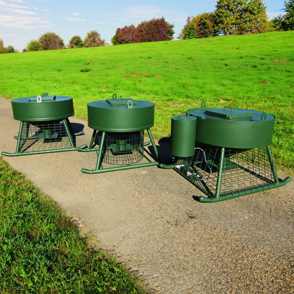 Automatic quail feeders placed on a pathway with a grassy background. The feeders are green with metal construction.