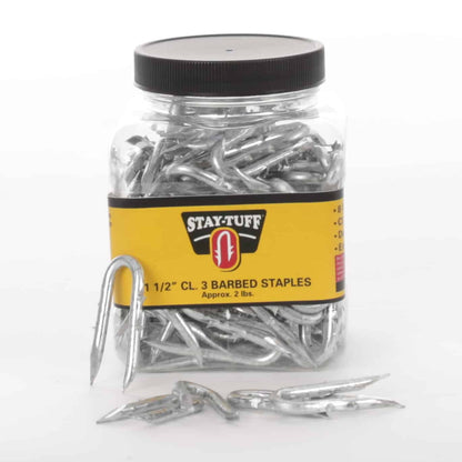 A clear plastic container with a black lid labeled 'STAY-TUFF' containing silver barbed staples, with some staples scattered outside the container.